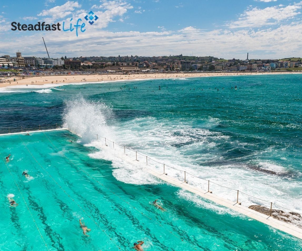 A beachfront scene welcomes you with a vibrant turquoise ocean and an outdoor swimming pool adjacent to the sea. Waves crash over the pool's edge as people swim. In the background, there is a sandy beach with visitors and a distant urban skyline. The text "Steadfast Life" is visible.