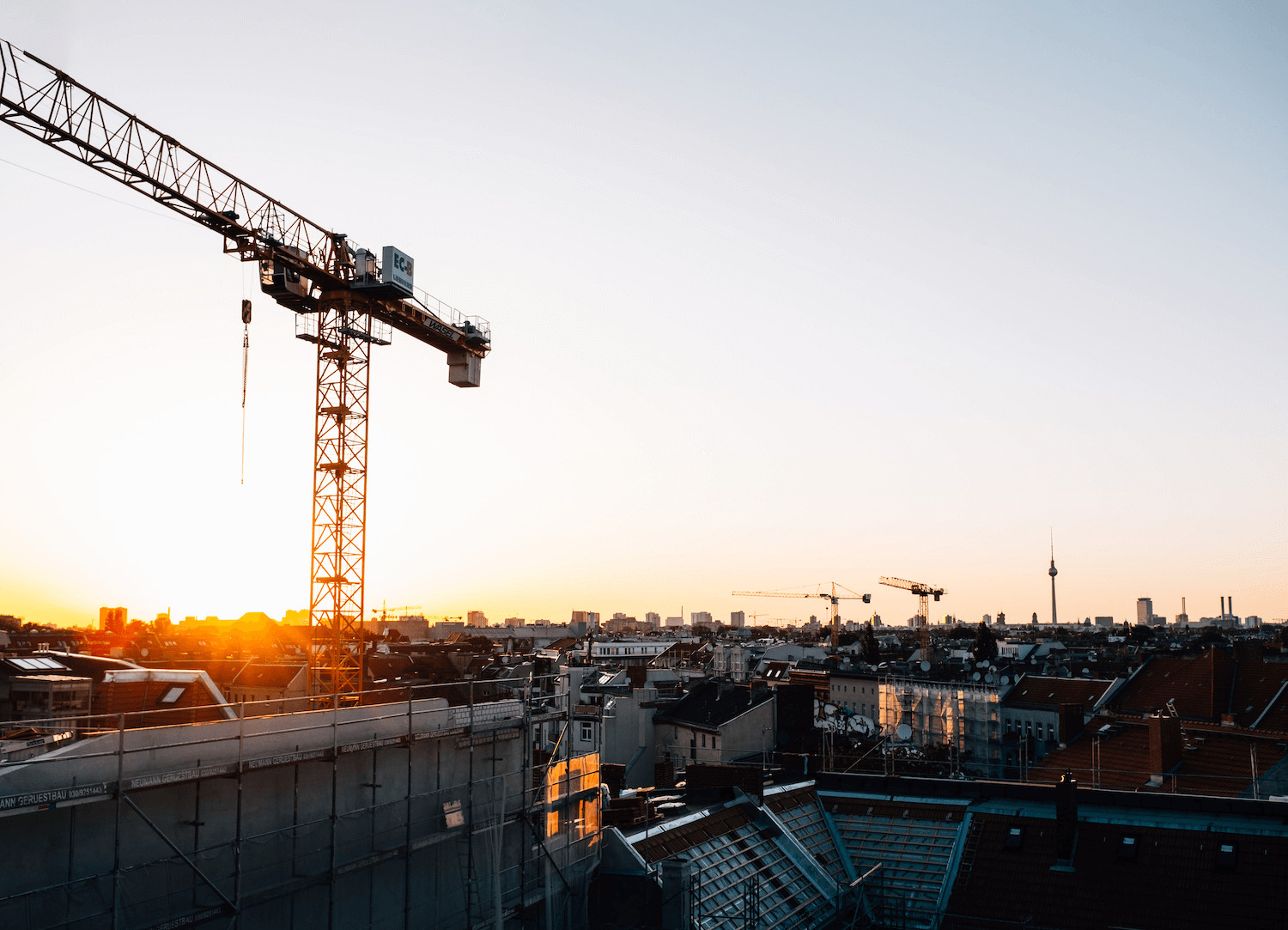 Cityscape at sunrise, featuring construction cranes and rooftops of buildings. The sky is clear with warm sunlight casting a glow over the urban area, highlighting a crane prominently to the left and the skyline in the background. It's a scene that underscores the importance of insurance for builders.