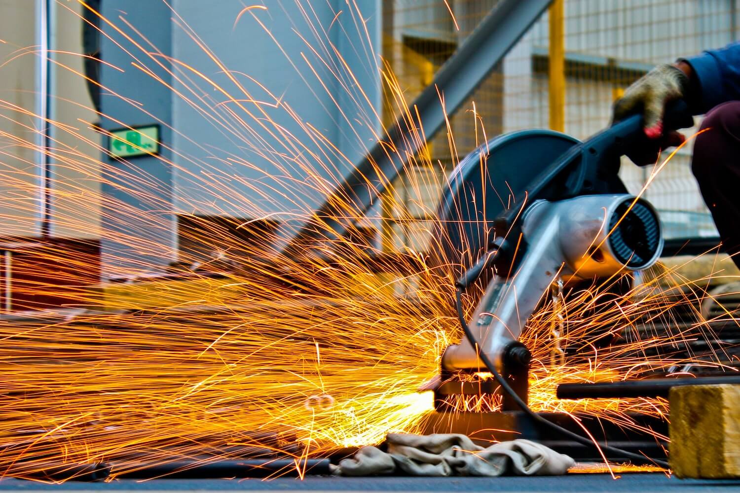 A worker operates a grinding machine, producing a shower of bright sparks. The worker wears protective gloves, focusing on the task. The background includes industrial equipment and safety barriers. This scene of intense manual labor underscores the importance of insurance for builders in such an industrial setting.