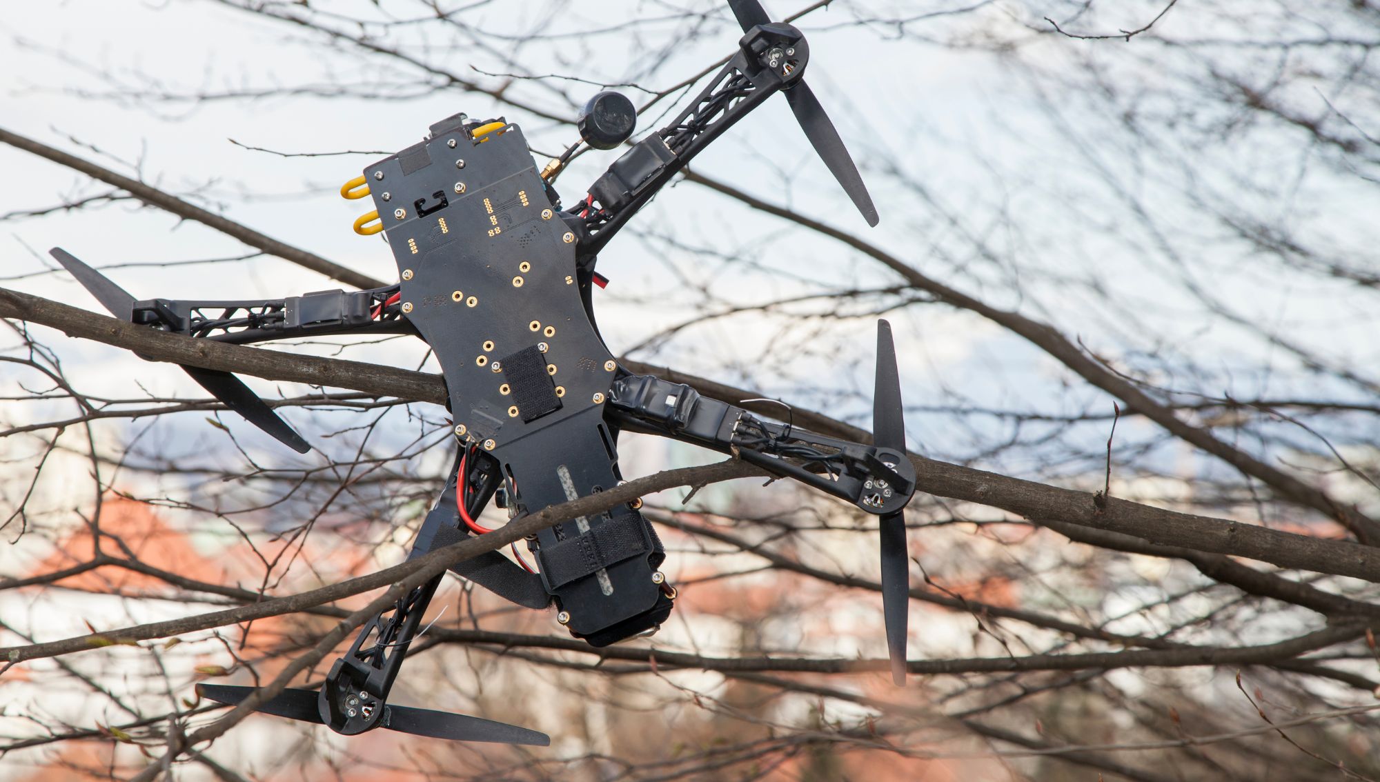 A black quadcopter drone is stuck in the branches of a tree. Its propellers are entangled among the branches, and it appears to be suspended, unable to fly. The background shows a blurred view of a town with houses and some natural landscape.