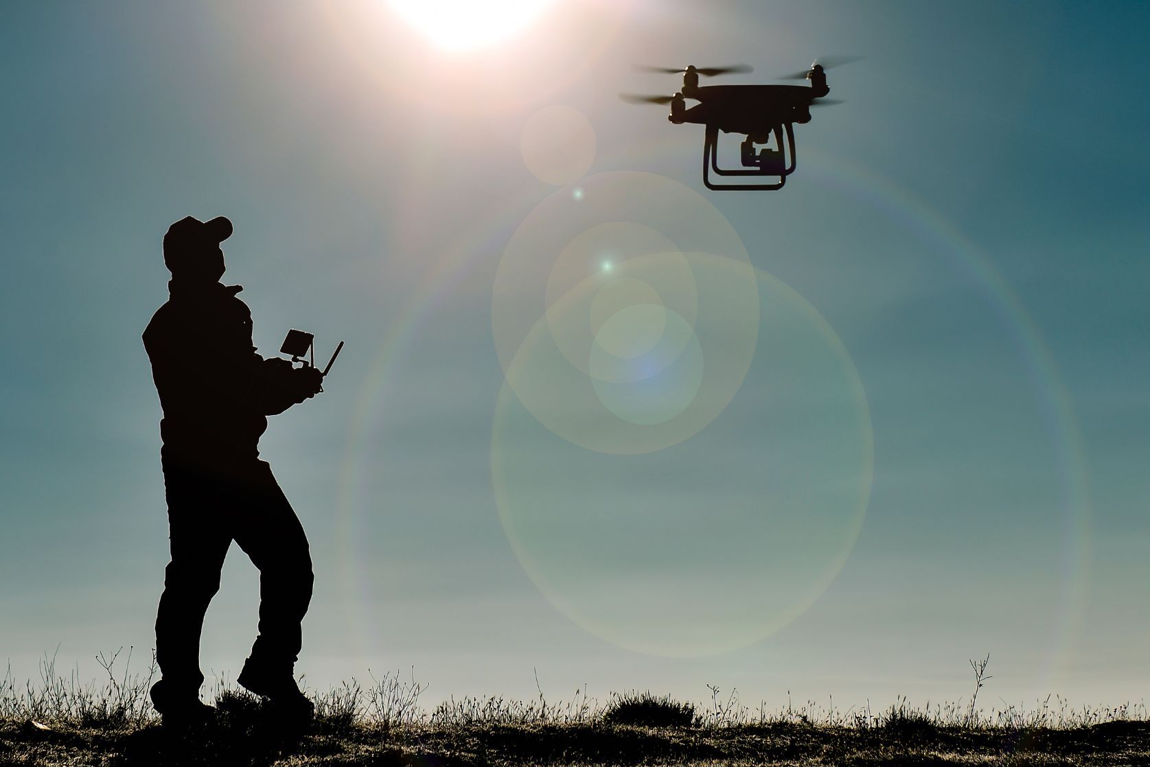 Silhouette of a person operating a drone with a remote control under a sunlit sky. The drone is in flight against a clear, bright background, casting lens flares. The person is standing on uneven ground, facing the drone.