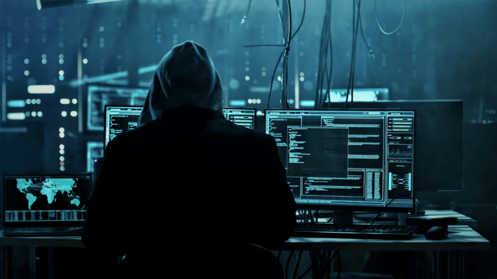 A person in a hoodie sits in front of multiple computer monitors displaying code and maps in a dark room, suggesting a hacking environment. Wires hang from the ceiling, enhancing the tech-centered atmosphere.