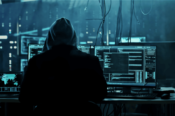 A person in a hoodie sits in front of multiple computer monitors displaying code and maps in a dark room, suggesting a hacking environment. Wires hang from the ceiling, enhancing the tech-centered atmosphere.